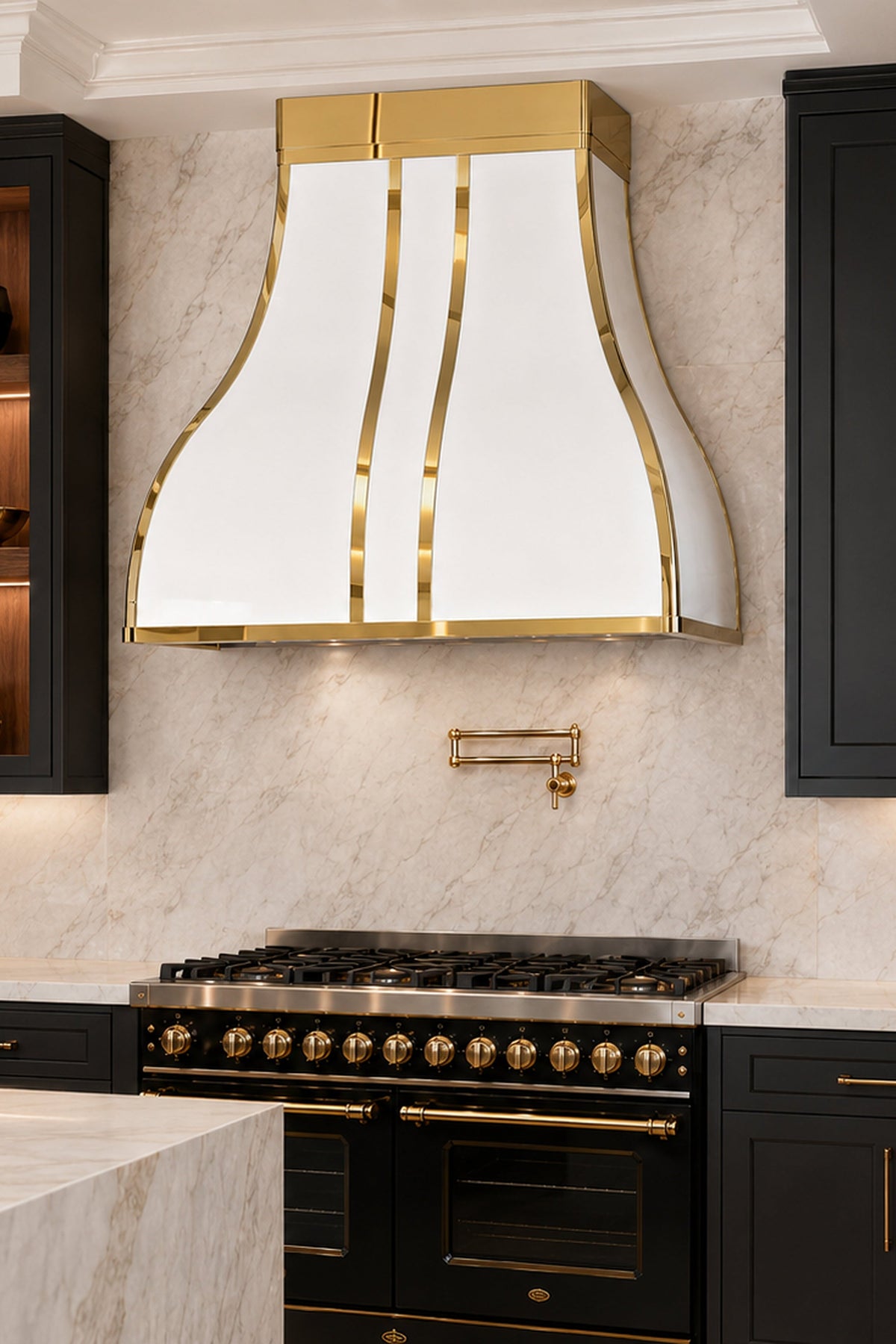 A luxury kitchen interior featuring a white and gold range hood mounted above a black gas range and oven, set against a light marble backsplash with black cabinetry and gold handles.
